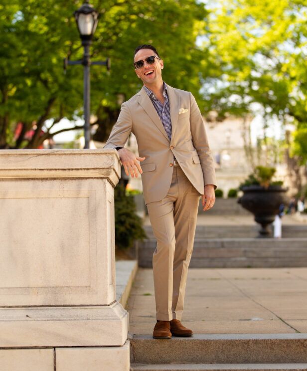 Khaki cotton suit with navy gingham shirt, cream pocket square and brown suede loafers, worn by a model outdoors