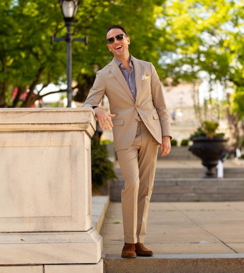 Khaki cotton suit with navy gingham shirt, cream pocket square and brown suede loafers, worn by a model outdoors