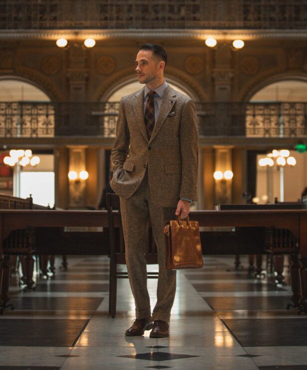 Brown Donegal tweed suit with blue striped shirt, plaid tie and brown leather briefcase, worn by a model in a library