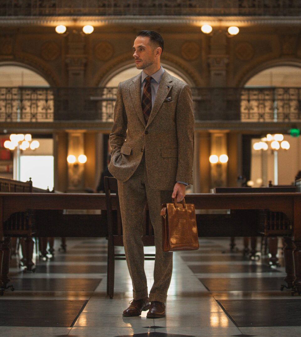 Brown Donegal tweed suit with blue striped shirt, plaid tie and brown leather briefcase, worn by a model in a library