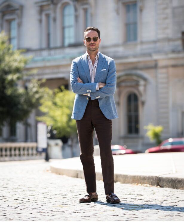 Light blue checked linen sport coat with striped shirt, brown trousers and brown loafers, worn by a model on a cobbled street