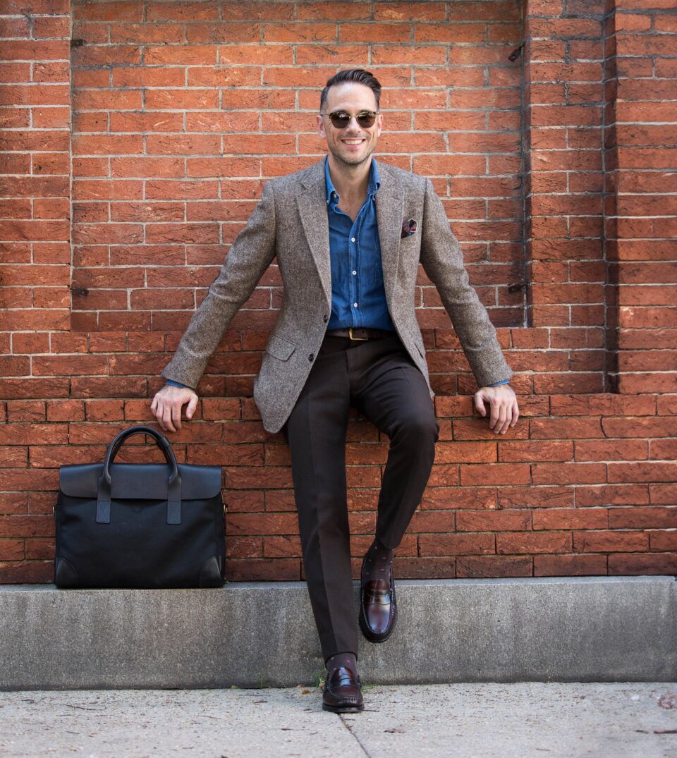 Brown donegal tweed sport coat with denim shirt, dark brown trousers and penny loafers, worn by a model against a brick wall