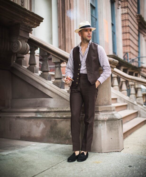 Brown linen waistcoat and matching trousers with gingham shirt, panama hat and black loafers, worn by a model on a stoop