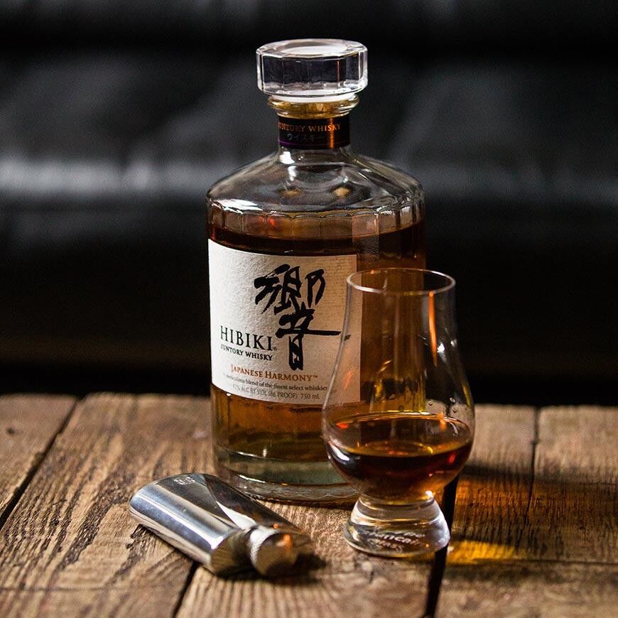 Polished pewter hip flask on rustic wood with a bottle of Hibiki Japanese whisky and filled tasting glass, styled scene