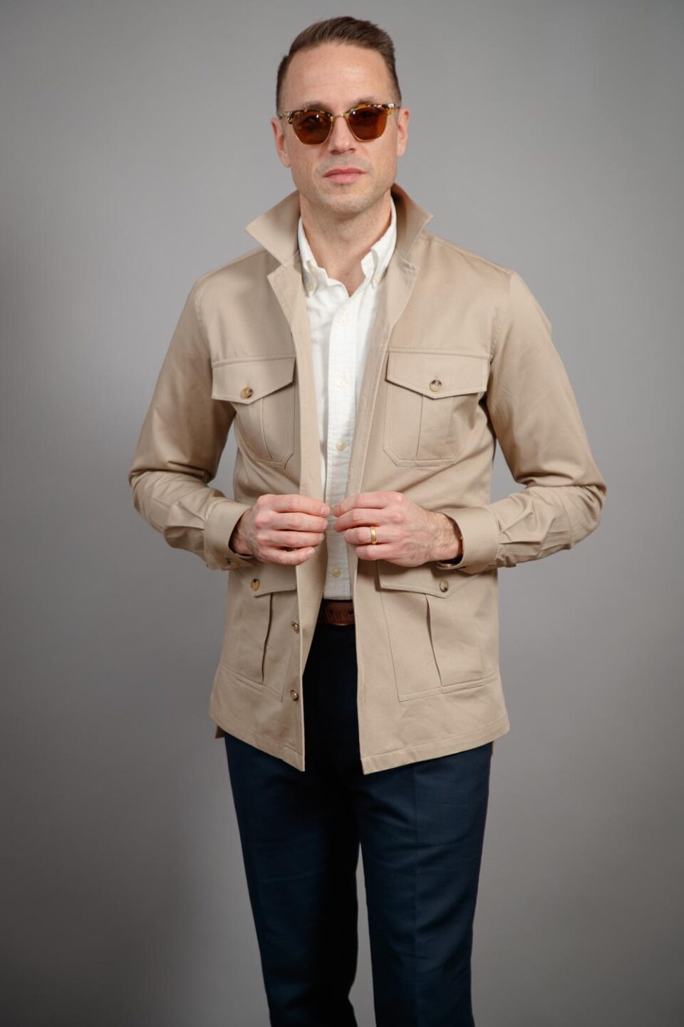 Tan cotton four-pocket safari jacket with stand collar, white shirt and navy trousers, worn by a model