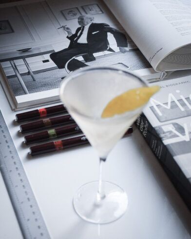 Martini with lemon twist beside an open Mies van der Rohe biography, pens and a ruler, in a styled desk scene
