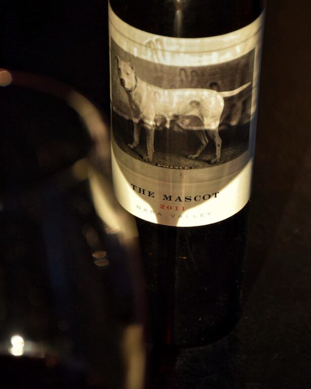 Bottle of 2011 The Mascot Napa Valley red wine with dog label, beside a filled glass in a dim styled scene