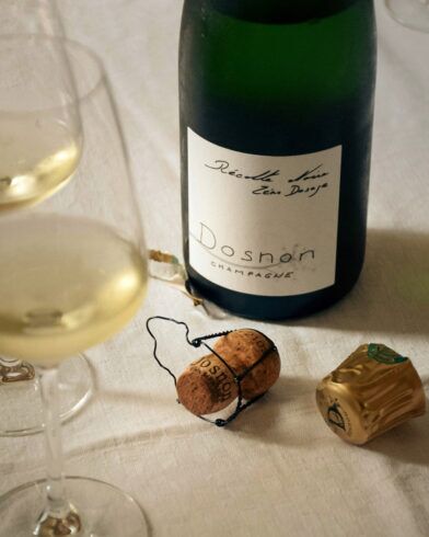 Open bottle of Dosnon champagne with poured glasses, cork and gold foil on a white linen table, a styled lifestyle scene