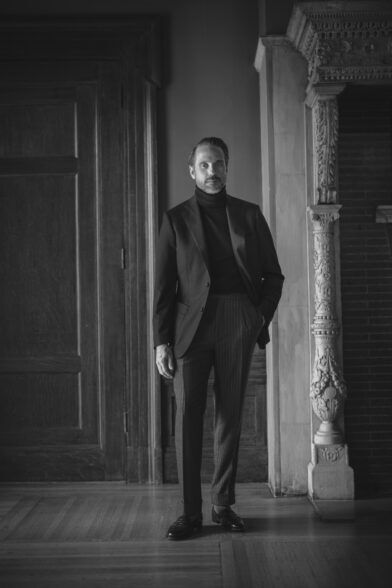 Black basket weave sport coat with dark turtleneck and pinstripe trousers, worn by a model in a stone hallway