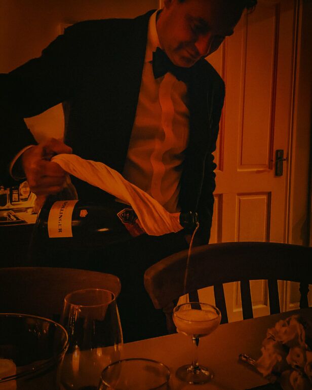 Black dinner jacket with white pleated shirt and black bow tie, worn by a man pouring champagne in a styled evening scene