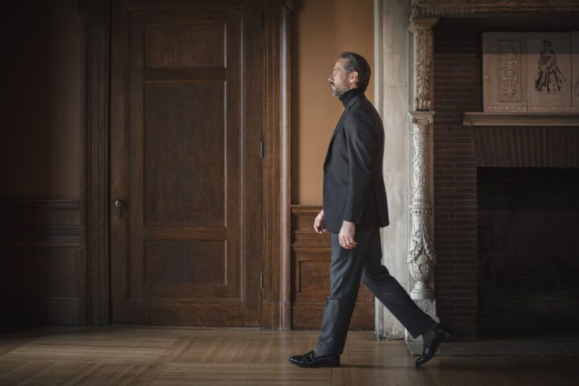 Black basket weave sport coat with black turtleneck, grey striped trousers and black loafers, worn by a model walking