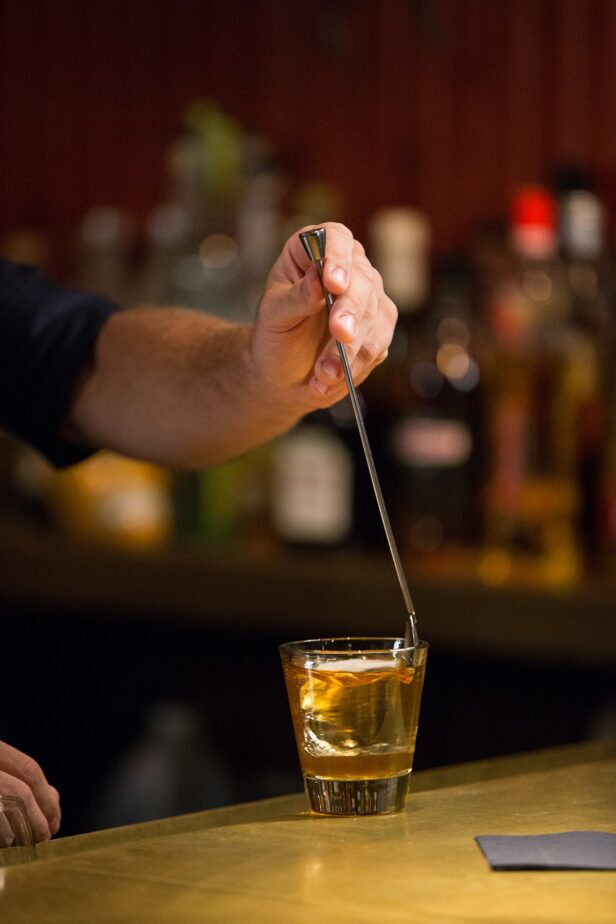 Old fashioned cocktail in a rocks glass being stirred with a bar spoon at a bar, a styled lifestyle scene