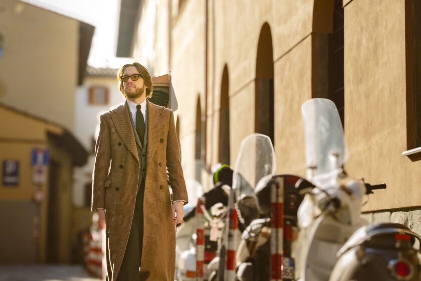 Tan glen check double-breasted overcoat with green waistcoat and black tie, worn by a model on a sunlit street