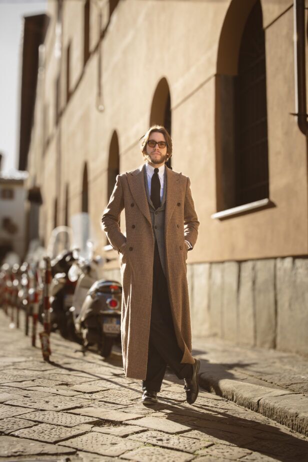 Brown glen check double-breasted wool overcoat with grey waistcoat and black tie, on a cobbled street