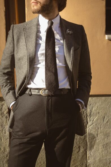 Charcoal grey flannel suit with white shirt, dark tie and lapel pin, worn by a model outdoors