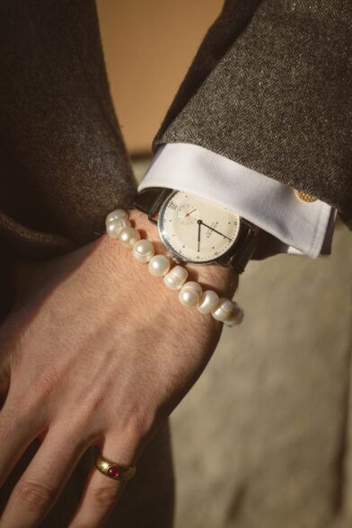 Baroque pearl bracelet and cream-dial watch with white shirt cuff and brown herringbone sleeve, a close-up detail