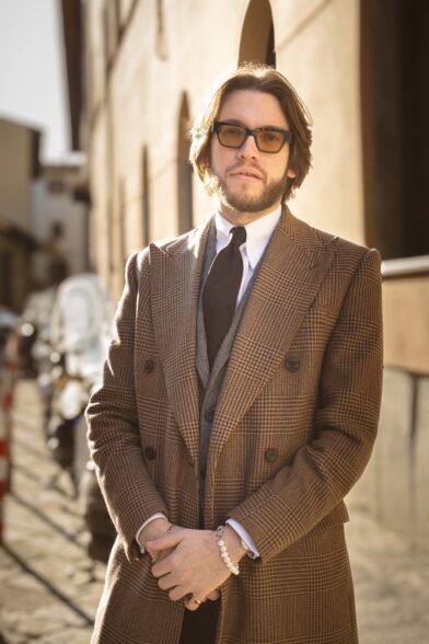 Brown glen check double-breasted wool overcoat with grey waistcoat and brown tie, on a sunlit street