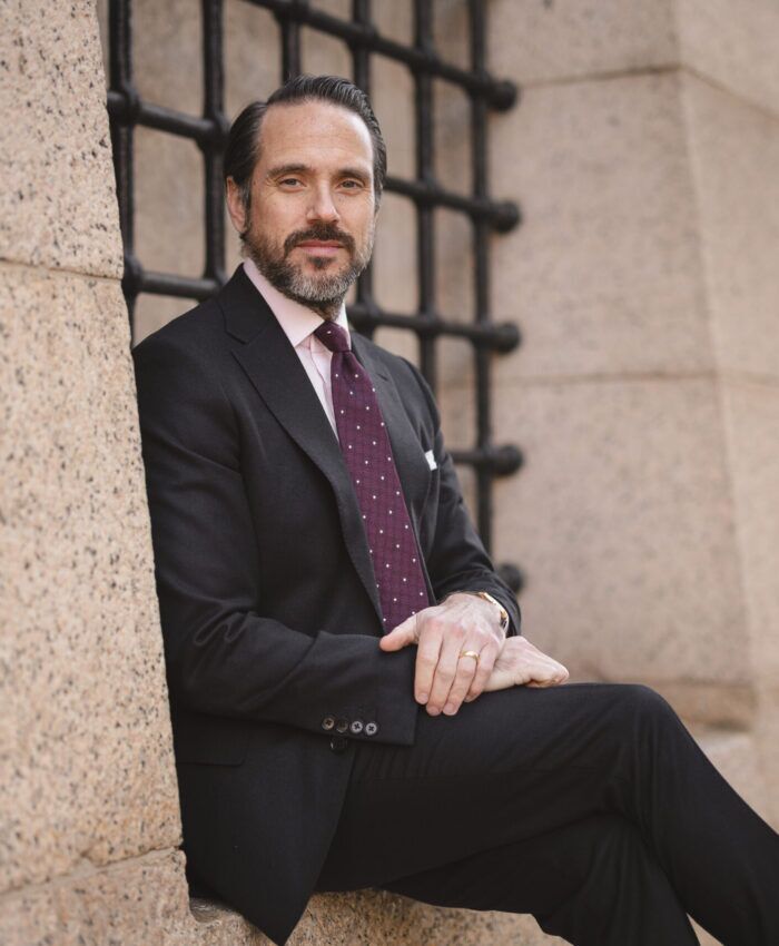 Black flannel suit with pink shirt, burgundy dotted silk tie and white pocket square, worn by a model seated outdoors