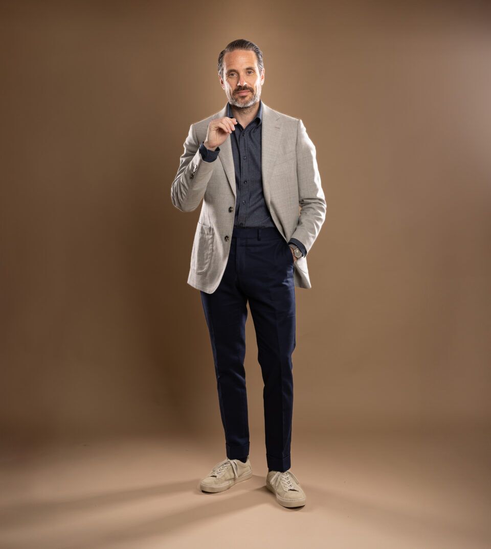 Light grey patch-pocket sport coat with navy denim shirt, navy trousers and taupe sneakers, worn by a model