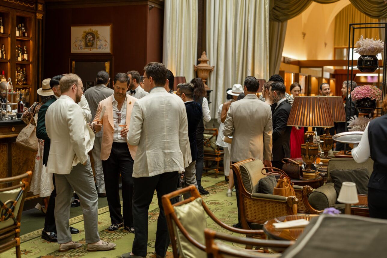 Peach linen sport coat with open white shirt and navy trousers, among cream summer tailoring at a hotel bar