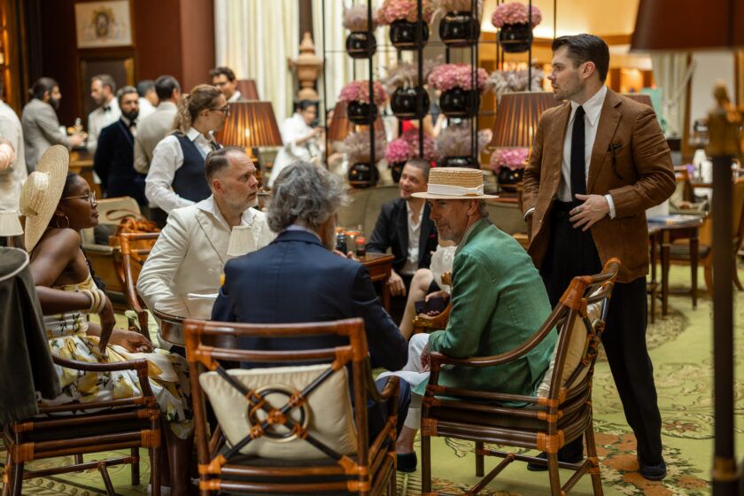 Tan linen sport coat with white shirt and black knit tie, among green linen and cream jackets in a hotel lounge scene