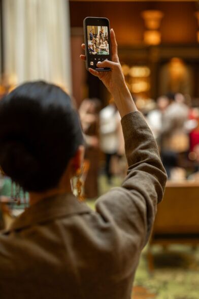 Textured brown jacket worn by a guest raising a phone to photograph a cocktail event crowd, a styled lifestyle scene