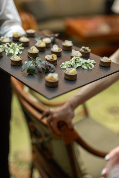 Dark wooden tray of canapés garnished with greenery, carried by a server at a cocktail event