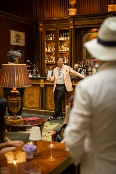 Peach linen sport coat with white shirt and charcoal trousers, worn by a man at a hotel bar in a styled lifestyle scene