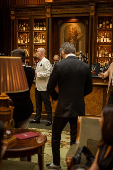Ivory dinner jacket with black trousers and velvet slippers, worn at a wood-panelled bar in a styled cocktail scene
