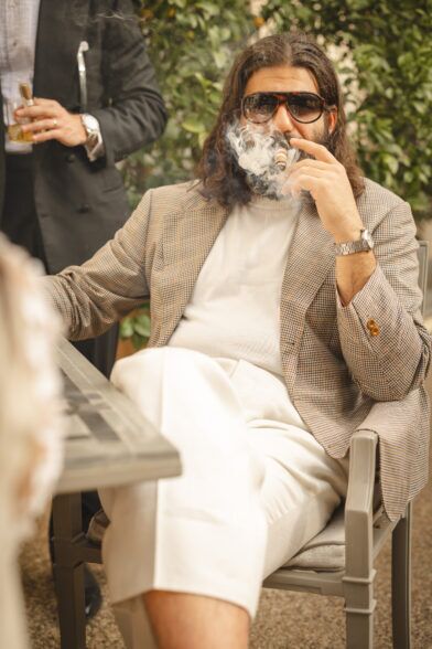 Tan and brown check sport coat with cream crewneck sweater and ivory trousers, worn by a seated man in a styled outdoor scene
