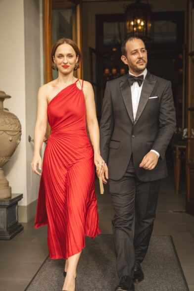 Charcoal peak lapel tuxedo with black bow tie and pocket square, worn beside a woman in a red gown