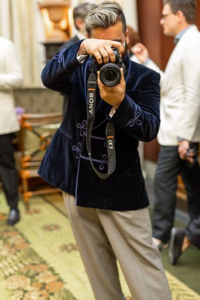Navy velvet smoking jacket with purple frogging and soutache trim, worn with grey trousers in a styled event scene