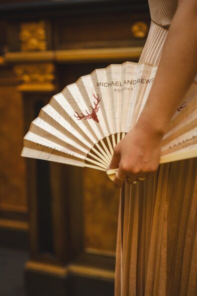 Cream paper hand fan printed with tailoring patterns and a red stag, held at an event, styled lifestyle scene