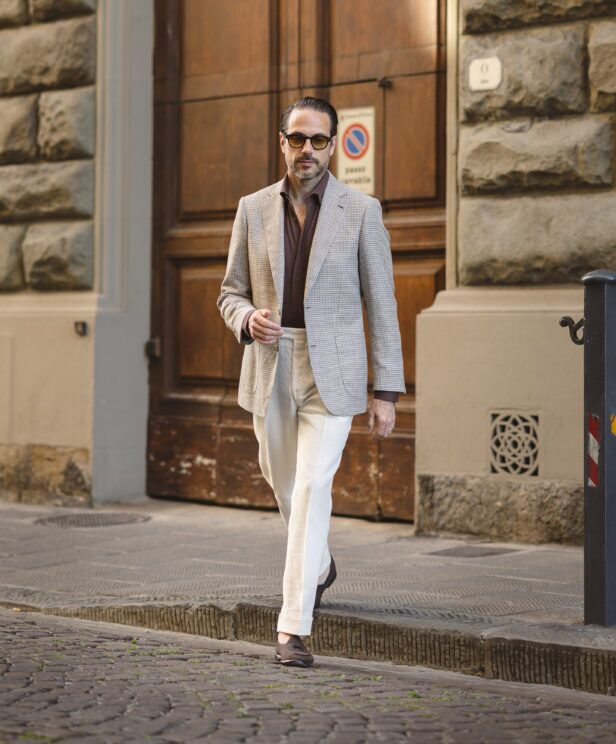 Tan and brown houndstooth sport coat with knit polo and cream trousers, worn on a cobbled street