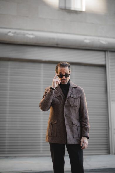 Brown flannel safari jacket over a black turtleneck, worn by a model adjusting sunglasses
