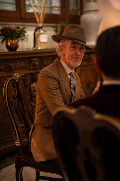 Tan wool jacket with a check tie and felt hat, worn by a seated guest