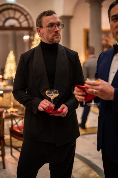 Black shawl collar dinner jacket over a turtleneck, worn by a guest holding a coupe