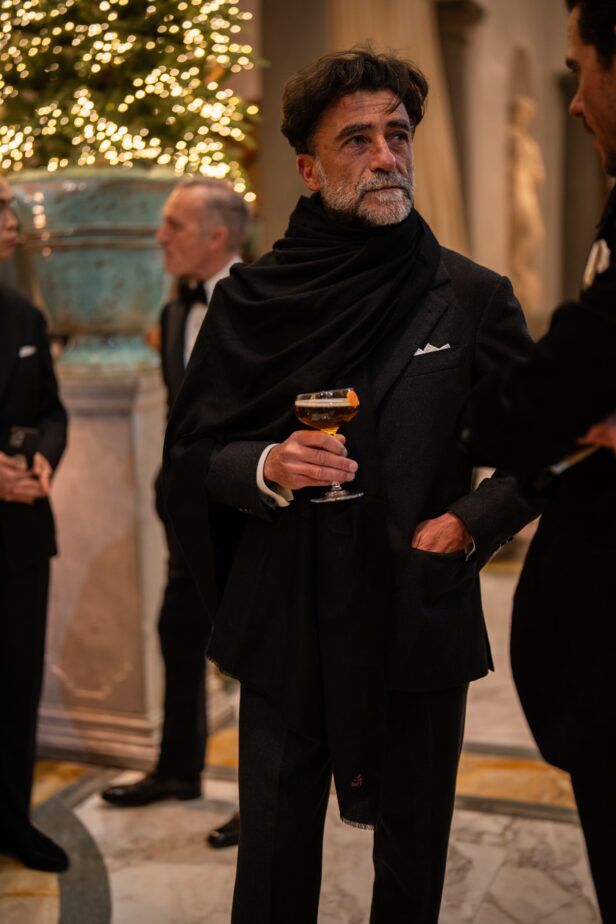 Black jacket with a wrapped black scarf and pocket square, worn by a guest with a drink