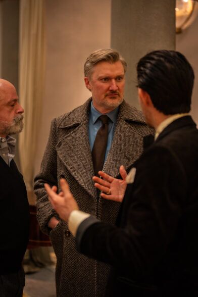 Grey herringbone tweed overcoat with a blue shirt and brown knit tie, worn at a party