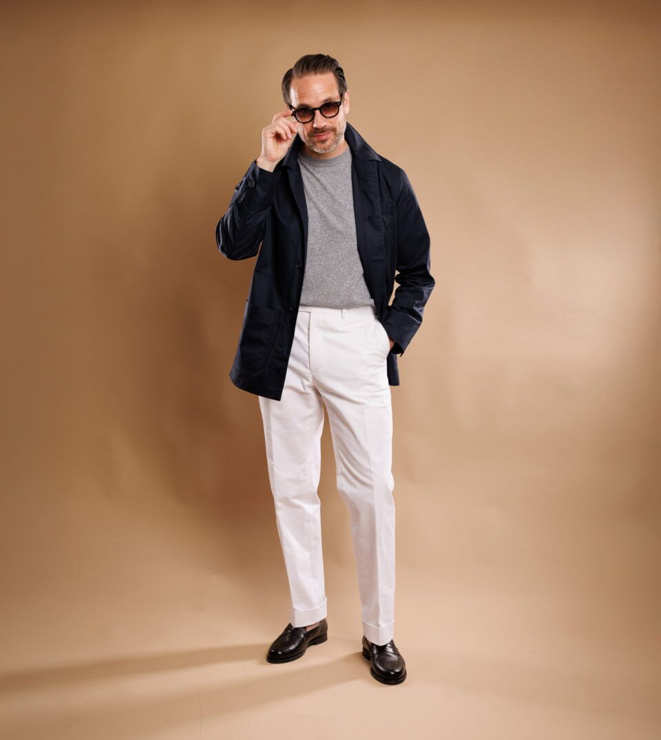 Navy twill chore coat with grey sweater, white trousers and brown loafers, full length