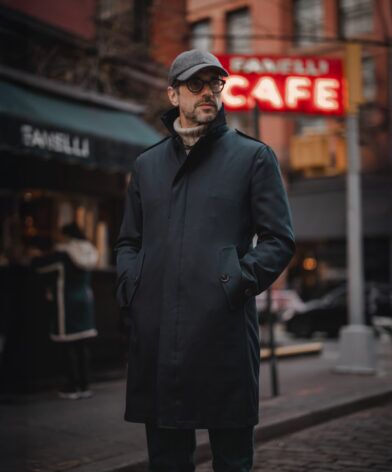 Navy storm coat over a cream turtleneck with a grey flat cap, worn by a model at night