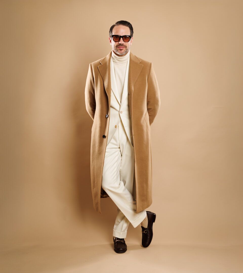 Camel overcoat over ivory flannel suit with cream turtleneck and brown bit loafers, worn by a model