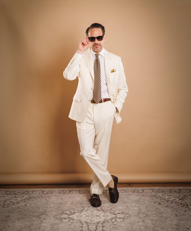 Ivory two-button suit with white shirt, brown geometric silk tie and brown bit loafers, worn by a model