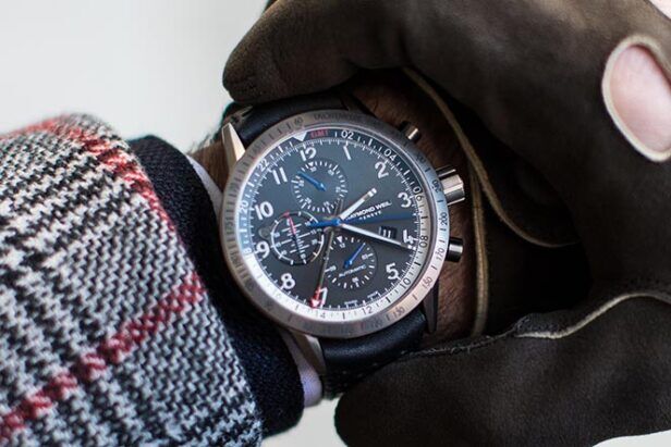 Black dial chronograph watch on black leather strap, worn with brown suede gloves and a checked coat sleeve, close-up