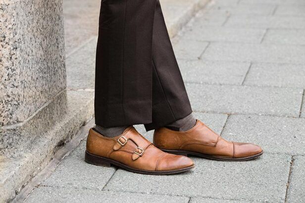 Tan leather double monk strap shoes with cap toes, worn with brown trousers and grey socks, a close-up detail