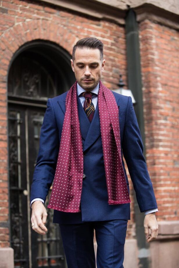 Burgundy silk scarf with white dots and fringed ends, worn with a navy three-piece suit by a model on a brick street
