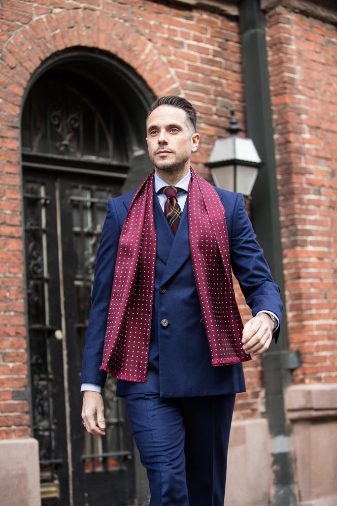 Burgundy silk scarf with white dots, worn with a navy three-piece suit and striped tie by a model on a city street