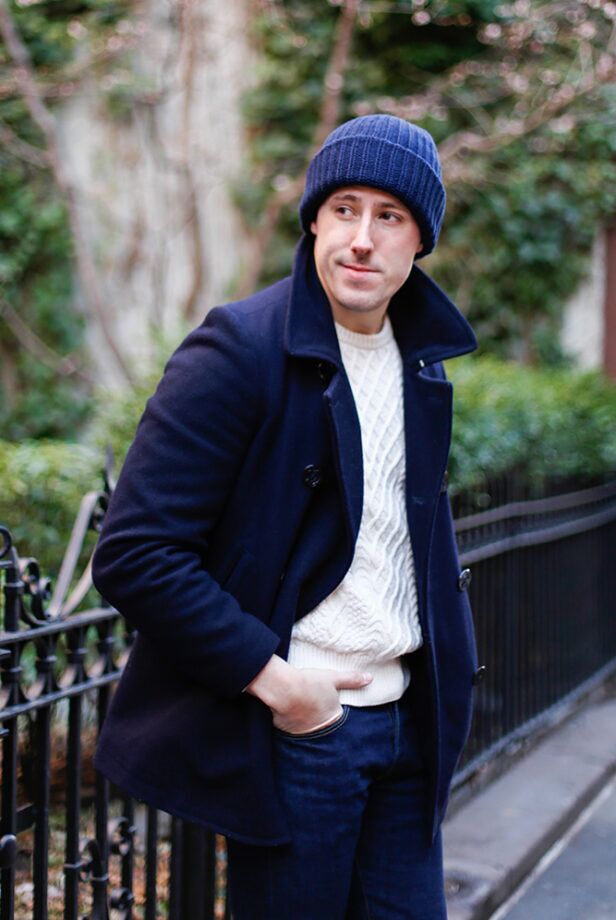 Navy wool peacoat with cream cable knit sweater, dark denim jeans and ribbed watch cap, worn by a model outdoors