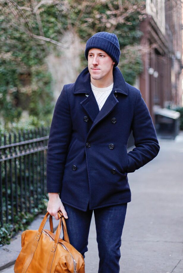 Navy wool double-breasted peacoat with cream cable knit sweater, dark denim and navy watch cap, worn by a model on a city street