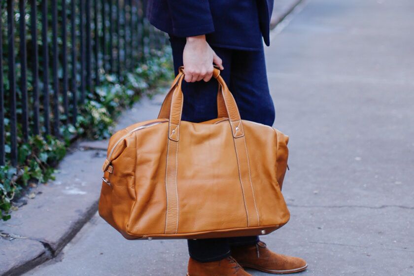 Tan leather weekender duffle bag with contrast stitching, carried by a model in navy denim and suede boots on a city street